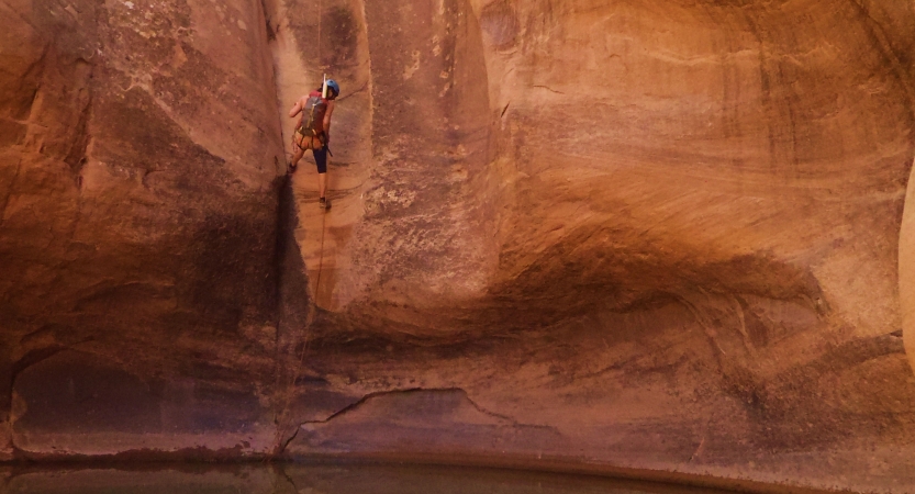 A person wearing safety gear is suspended by ropes mid air against the backdrop of a red canyon wall.